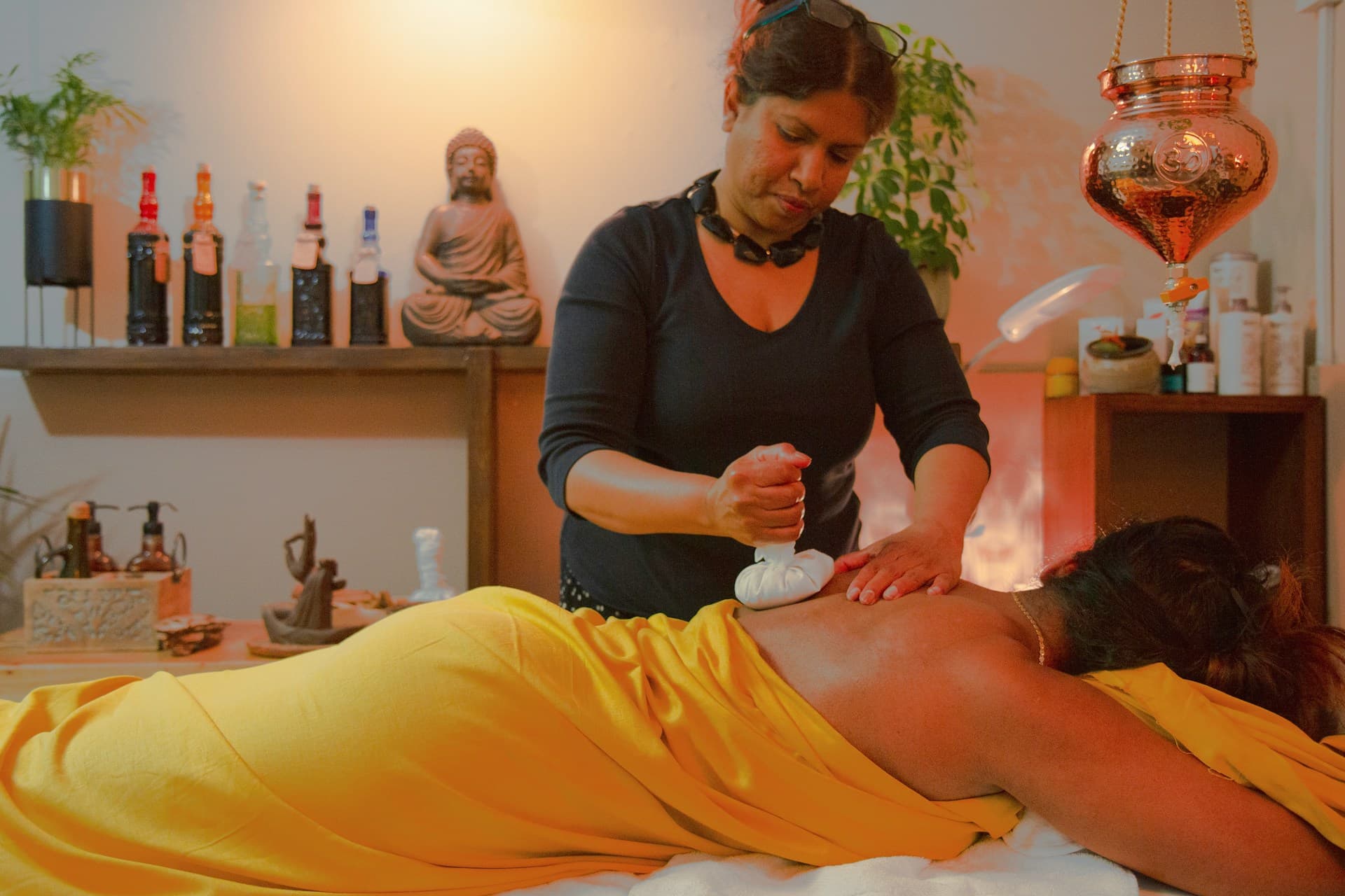 herapist performing an Ayurvedic-style Abhyanga oil massage using a warm herbal compress on a client’s back in a calm treatment room.